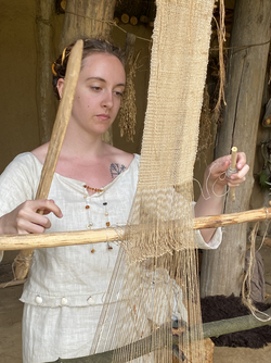 Frau in weißem Leinenkleid webt an einem senkrechten Webstuhl aus Holz