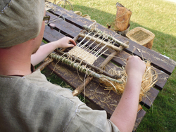 Person in historischer Kleidung sitzt an einem Holztisch und webt mit einem einfachen Webrahmen aus Holz