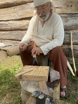 Person in historischer Kleidung sitzt auf Holzscheit und bearbeitet Holz mit Meißel und Holzhammer vor Holzwand