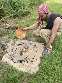 Eine Person mit rot-weiß kariertem Kopftuch und ärmellosem Oberteil kniet auf Gras und schüttet Haselnüsse aus einem flachen Korb auf eine Sandfläche mit Holzkohleresten