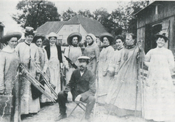 Schwarz-weiß-Fotografie einer Gruppe von Frauen in langen Kleidern und Hüten, eine sitzende Person in Anzug, im Hintergrund ein großes Haus und Bäume