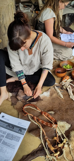 Person sitzt auf dem Boden und bearbeitet ein Lederstück mit mehreren Leder- und Fellstücken sowie einem geflochtenen Lederriemen vor sich